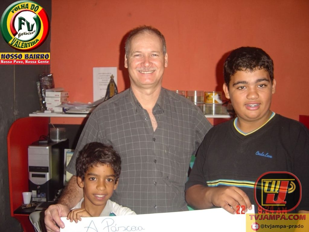 Fotografia de 2008 em um escritório de jogos, Byte Games, em João Pessoa, Paraíba. O jornalista Petrucio Prado, representando a Folha do Valentina (FV) e a TV JAMPA Oficial Jornal Jampa Notícias, sorri ao lado de dois jovens. Os três seguram um cartaz de cartolina branco com a escrita manuscrita 'A Páscoa'. O adolescente à direita, o outro de camisa preta com listras coloridas, segura uma das extremidades do cartaz. O menino mais novo à esquerda também segura, sorrindo. O fundo mostra estantes de livros, uma torre de PC e uma pequena estátua religiosa de Nossa Senhora de Aparecida. Uma data digital vermelha '22 9:24 AM' está no canto inferior direito.