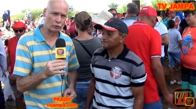 Registro em vídeo do arquivo Compositor JJ do Nordeste.JPG durante o programa Jampa Notícias, mostrando o repórter Petrúcio Prado vestindo camisa polo listrada em azul e amarelo e segurando o microfone da TV JAMPA, enquanto entrevista o compositor JJ, que veste uma camisa listrada em cinza, preto e branco com boné, em meio ao público reunido na cidade de Monteiro, Paraíba.