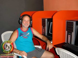 Petrúcio Prado TV JAMPA oficial sentado em uma estação da Byte Games Lan House, usando headset e sorrindo para a câmera durante uma pausa no trabalho. Ao fundo, aparecem computadores e cabines de acesso à internet que marcaram uma época no bairro Valentina.
