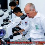 “Professor e biomédico Petrúcio Prado utilizando microscópio em laboratório de Biomedicina, acompanhado de estudantes durante atividade prática científica. A imagem mostra equipamentos laboratoriais e ambiente acadêmico de pesquisa, com destaque para o jaleco branco identificado com o nome Petrúcio Prado e a inscrição ‘BIOMEDICINA’ em verde. Na parte inferior aparece a marca Jornal Jampa Notícias – TV Jampa.”