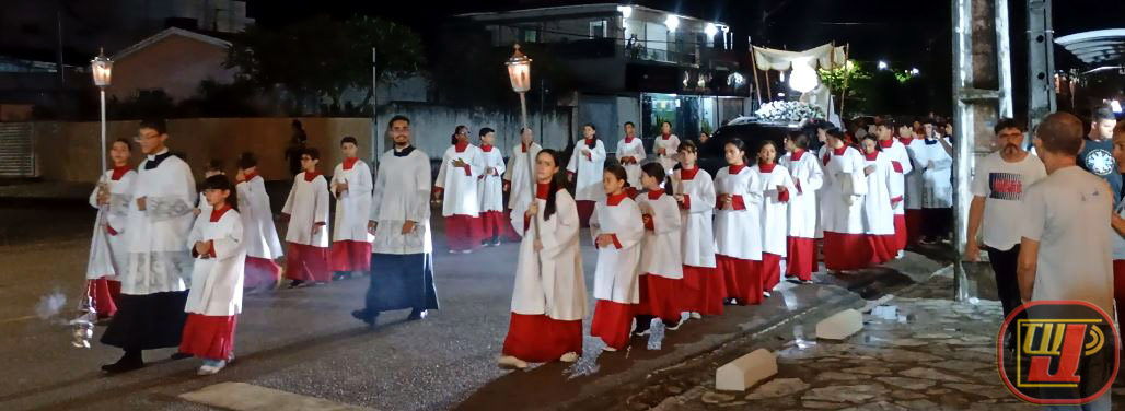 Fotografia noturna mostrando a procissão de Corpus Christi pelas ruas do bairro Valentina Figueiredo. Em destaque, um grande grupo de coroinhas e acólitos vestindo túnicas brancas e debruns vermelhos caminha em cortejo organizada pelo asfalto. Alguns seguram tochas acesas e um turíbulo com incenso. Ao fundo, vê-se o dossel que protege o Santíssimo Sacramento e moradores acompanhando nas calçadas. No canto inferior direito, há o logotipo da TV Jampa.