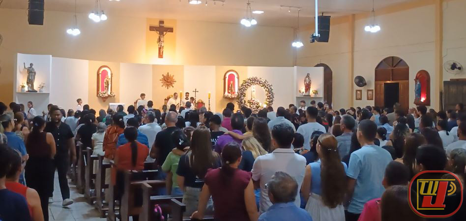 Um registro fotográfico de uma igreja lotada de fiéis durante uma celebração religiosa. A imagem, intitulada 31-05-2026 - Coroação Nossa Senhora - Paróquia aparecida - TV JAMPA Prado 2.jpg, mostra a assembleia de costas, voltada para o altar ao fundo. O altar está ornamentado com a cruz central, várias imagens de santos, incluindo uma imagem proeminente de Nossa Senhora, e um grande arranjo floral. Várias pessoas participam da celebração no presbitério. O templo tem iluminação suspensa e janelas em arco na lateral. No canto inferior direito, há uma logomarca "TV J" vermelha e amarela.