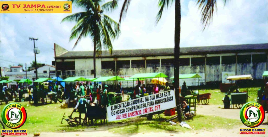 : Fotografia colorida capturada em plano médio que destaca a movimentação na feira livre ao lado do Mercado Público do Valentina. Em primeiro plano, uma banca exibe frutas frescas organizadas, com bananas em destaque. Ao fundo, feirantes e clientes interagem sob coberturas e tendas comerciais. A cena possui iluminação natural do dia, registrando a rotina e o comércio popular local.