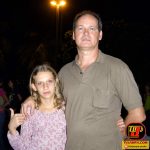 Uma fotografia colorida tirada à noite em um ambiente ao ar livre. À direita, um homem adulto de pele clara, cabelos curtos castanhos e bigode veste uma camisa polo marrom. Ele está com o braço direito apoiado no ombro de uma jovem garota que está à sua esquerda. A menina, de pele clara e longos cabelos loiros ondulados, veste uma blusa estampada em tons de rosa e branco e faz um sinal de "positivo" com o polegar direito. Ao fundo, vê-se a iluminação de um poste de luz forte e silhuetas de árvores e pessoas. No canto inferior direito, há uma marca d'água com a logo da TV Jampa. Petrucio Prado e ua filha Amanda Prado