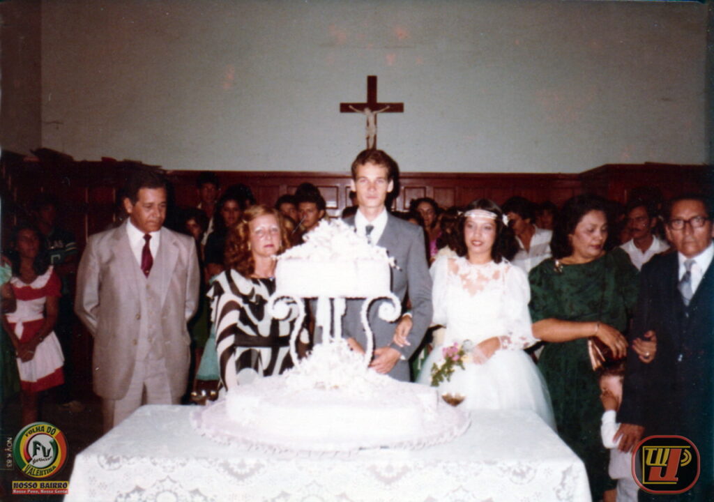 Fotografia histórica com cores restauradas do casamento de Petrúcio Prado e Rejane Moreira Prado, realizada em 8 de outubro de 1983. O casal aparece posicionado ao centro, atrás de um grande bolo de casamento decorado sobre uma mesa com toalha de renda branca. Petrúcio veste terno cinza com gravata e Rejane usa vestido de noiva clássico com detalhes em renda e tiara. Familiares e convidados posam ao lado e ao fundo em um ambiente solene com um crucifixo na parede. No canto inferior esquerdo constam as marcas "Folha do Valentina / Nosso Bairro" e, no canto inferior direito, o logotipo antigo da TV Jampa.