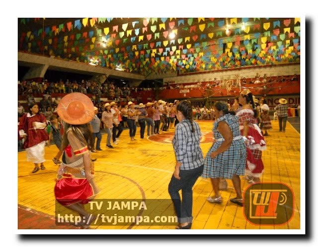 Apresentação de quadrilha junina em uma quadra coberta e decorada com bandeirinhas coloridas, mostrando pessoas com trajes caipiras dançando durante o evento do Projeto Vida Saudável no Valentina.