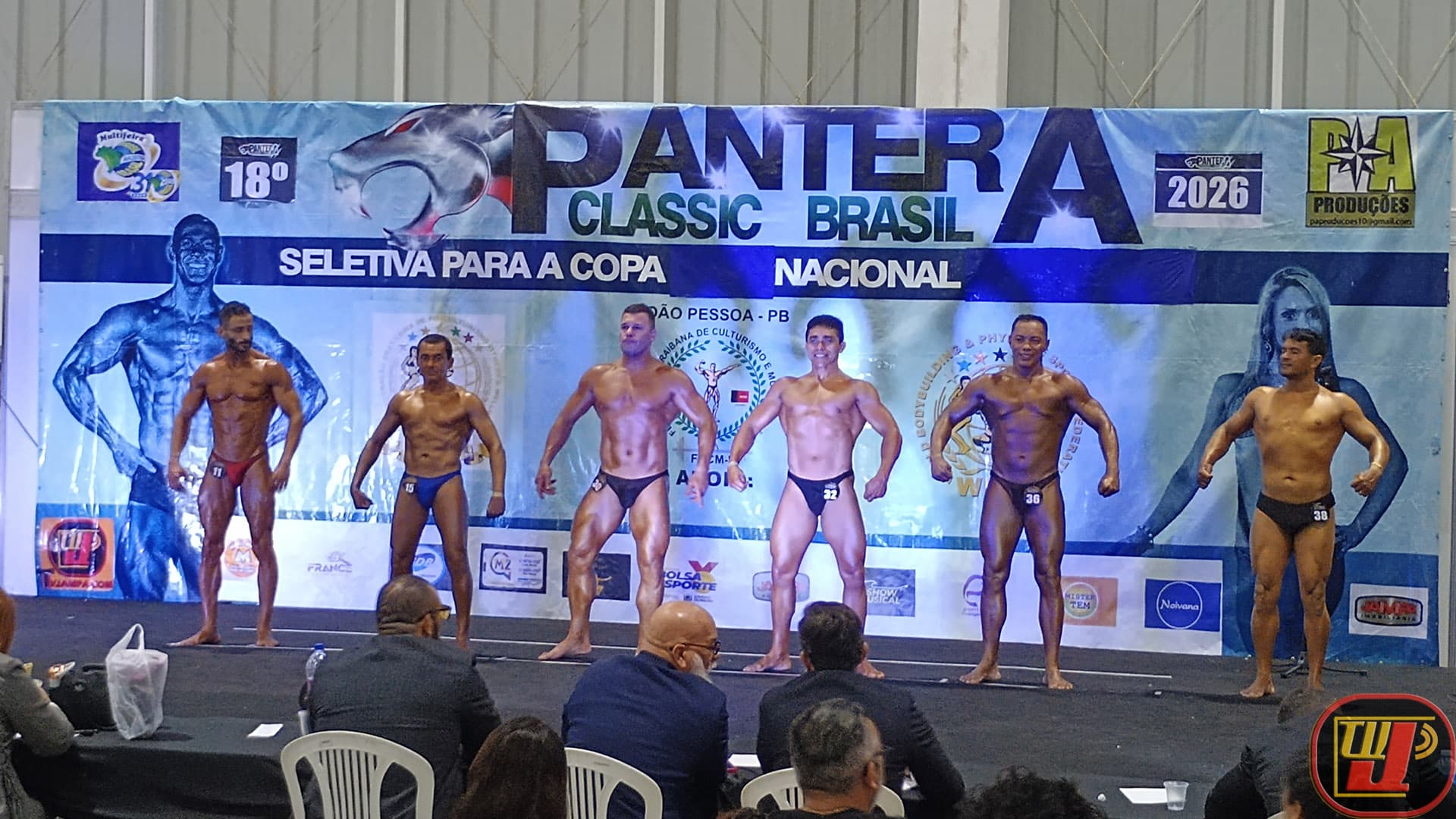 Seis atletas de fisiculturismo masculino posando em linha no palco durante o 18º Campeonato Pantera Classic Brasil 2026, com o painel oficial ao fundo e a mesa dos árbitros em primeiro plano.