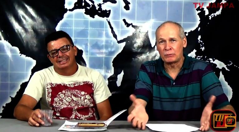 Dois homens sentados lado a lado atrás de uma bancada de estúdio em uma gravação da TV Jampa. À esquerda Cláudio Tel Pastel e à direita, o apresentador Petrucio Prado fala gesticulando.