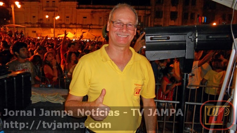 Homem sorridente de óculos e camisa polo amarela faz um gesto de positivo com o polegar. Ele está em primeiro plano, posicionado em um evento noturno ao ar livre no Ponto de Cem Réis. Ao fundo, uma grande multidão se aglomera perto de prédios históricos iluminados. À direita, há um equipamento de iluminação de palco. Na parte inferior da imagem, há uma faixa translúcida com o texto "Jornal Jampa Notícias - TV JAMPA" e o site "http://tvjampa.com", além do logotipo da TV Jampa no canto inferior direito.