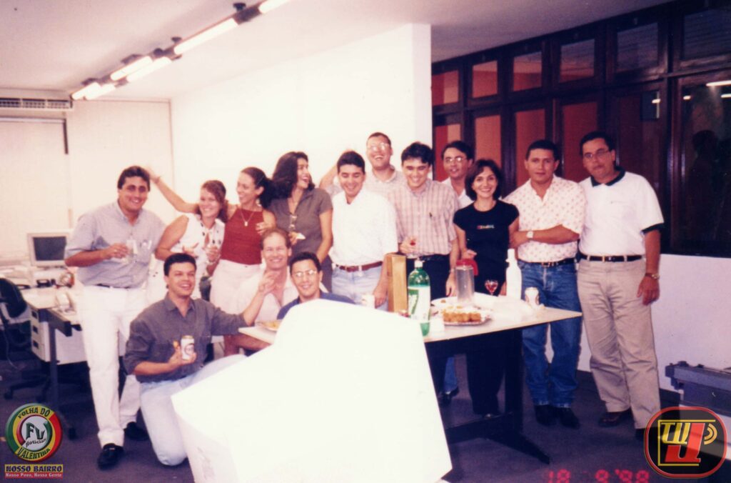 Fotografia colorida de setembro de 1998 mostrando uma confraternização interna de funcionários no escritório do Banco ABN AMRO BANK. Cerca de quatorze pessoas posam sorridentes ao redor de uma mesa com bolos, garrafas de refrigerante e copos. Alguns funcionários estão de pé e outros agachados ou sentados. O ambiente de escritório possui computadores antigos de tubo ao fundo, divisórias brancas e janelas de vidro com moldura escura. No canto inferior esquerdo, há o logotipo "Folha do Valentina / Nosso Bairro" e, no canto direito, o logotipo "TV Jampa". No canto inferior direito, a data gravada em vermelho registra "18 9 '98".