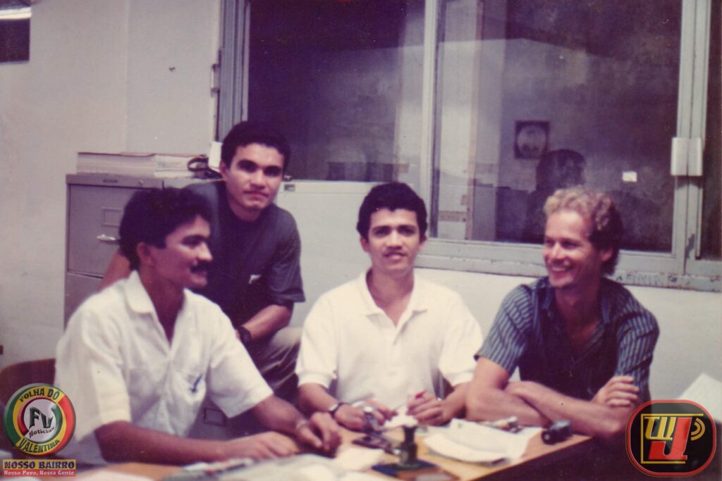 Foto histórica em ambiente interno, registrada no Banco Bandeirantes em 1987. Quatro homens jovens estão reunidos ao redor de uma mesa de escritório de madeira ao final do expediente. Três deles estão sentados e um permanece de pé ao fundo. Eles sorriem e conversam em um momento de descontração. No canto inferior esquerdo constam as marcas "Folha do Valentina" e "Nosso Bairro", e no canto inferior direito, o logotipo antigo.