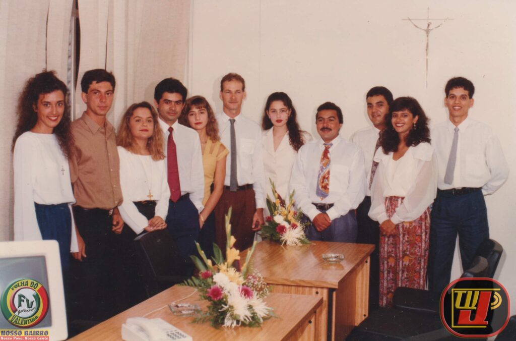 Foto antiga da equipe do Banco Bandeirantes S.A. em Campina Grande, com dez funcionários alinhados em pé atrás de mesas de escritório de madeira com arranjos de flores.