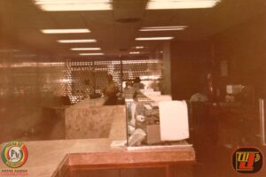 Fotografia tirada por Petrúcio Prado mostrando funcionário do Banco Bandeirantes trabalhando em máquina de datilografia nos anos 80 em João Pessoa, cercado por papéis e colegas de escritório.