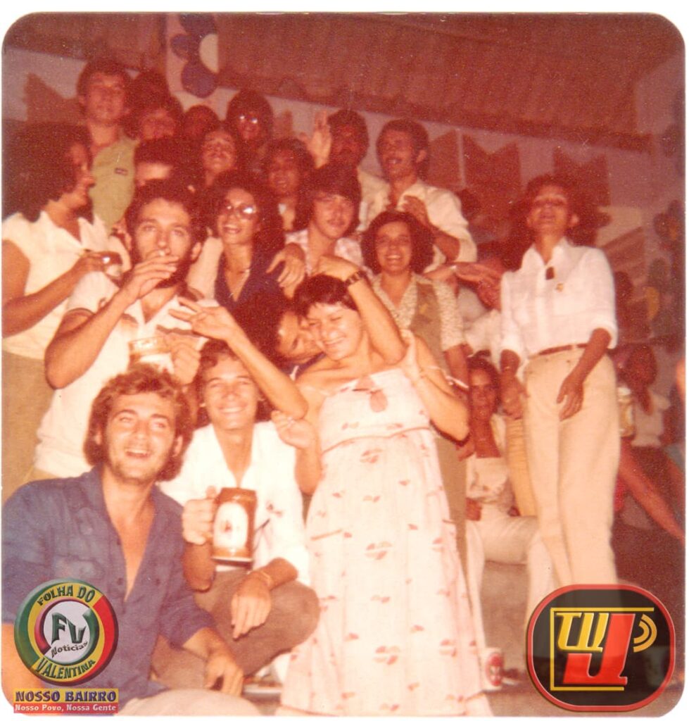Foto histórica com tom envelhecido de uma grande confraternização em grupo em ambiente festivo. No canto inferior esquerdo, o jovem Petrucio Prado aparece agachado vestindo camisa branca e segurando uma caneca escura com estampa clara. Ao lado dele, outros jovens posam sorrindo, incluindo um homem de camisa azul na frente e uma mulher grávida com vestido estampado tomara-que-caia logo atrás. Diversas pessoas estão agrupadas em níveis diferentes ao fundo, celebrando e acenando sob uma estrutura com teto de telhas. No canto inferior esquerdo, há o logotipo da "Folha do Valentina" e, no canto inferior direito, o logotipo da TV Jampa.