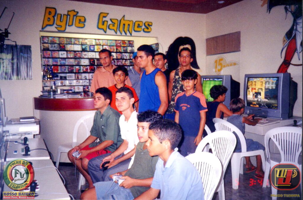 TEXTO ALTERNATIVO (Acessibilidade / Alt Text)Foto de 2005 em plano médio mostrando o interior de uma locadora de videogames chamada "Byte Games", com cerca de quinze jovens e crianças. No centro e à direita, garotos jogam sentados em cadeiras de plástico brancas em frente a televisores de tubo antigos. Outros jovens em pé aguardam a vez ao redor do balcão de atendimento, que possui uma prateleira cheia de mídias de jogos ao fundo. No canto inferior esquerdo, há um computador com o logotipo do jornal "Folha do Valentina". No canto inferior direito, o logotipo "TVJAMPA".