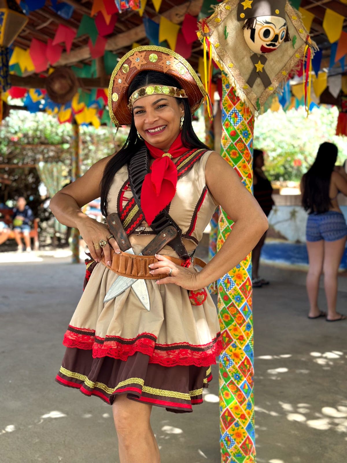 A cantora Ana Virgínia sorri para a foto em um galpão rústico decorado com bandeirinhas e enfeites juninos coloridos. Ela está caracterizada como Maria Bonita, usando chapéu de couro ornamentado, lenço vermelho no pescoço, roupa típica em tons de bege e marrom com detalhes vermelhos e dourados, e uma cartucheira com peixeiras artesanais cruzadas na cintura.
