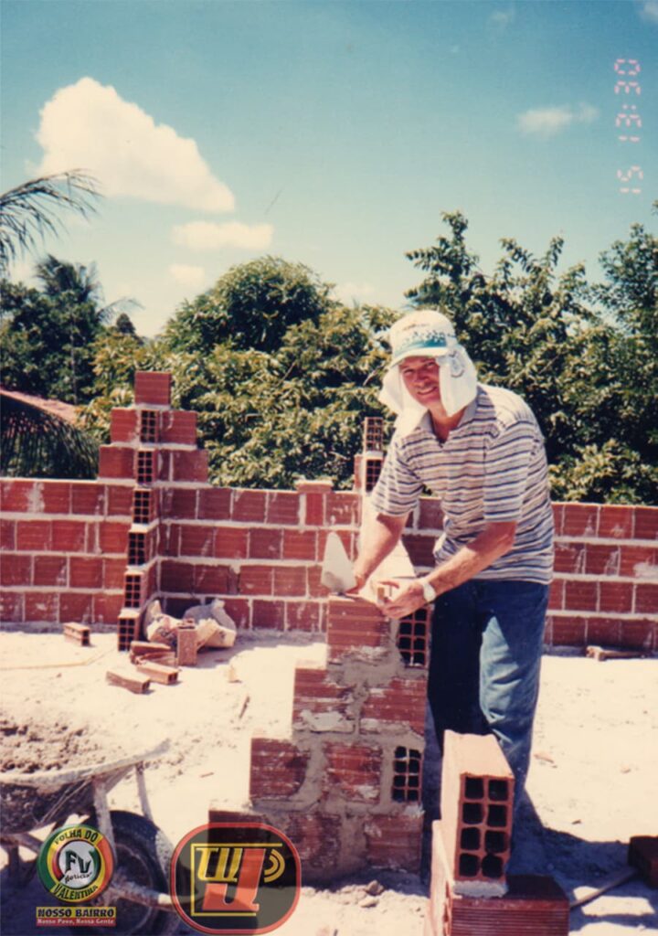Fotografia histórica em ambiente externo e sob a luz do dia, mostrando Petrúcio Prado trabalhando na fase inicial da construção de uma parede de tijolos. Ele veste uma camisa polo listrada, calça jeans e um chapéu branco com proteção de nuca para o sol. Com uma colher de pedreiro na mão direita, ele assenta a massa sobre os tijolos cerâmicos. Ao fundo, vê-se a copa de árvores verdes sob um céu azul com nuvens claras. No canto inferior esquerdo está a marca circular "Folha do Valentina Notícias" e, na parte inferior central, o logotipo vermelho e amarelo da TV Jampa. No canto superior direito consta a marcação de data em vermelho "15 13:30".