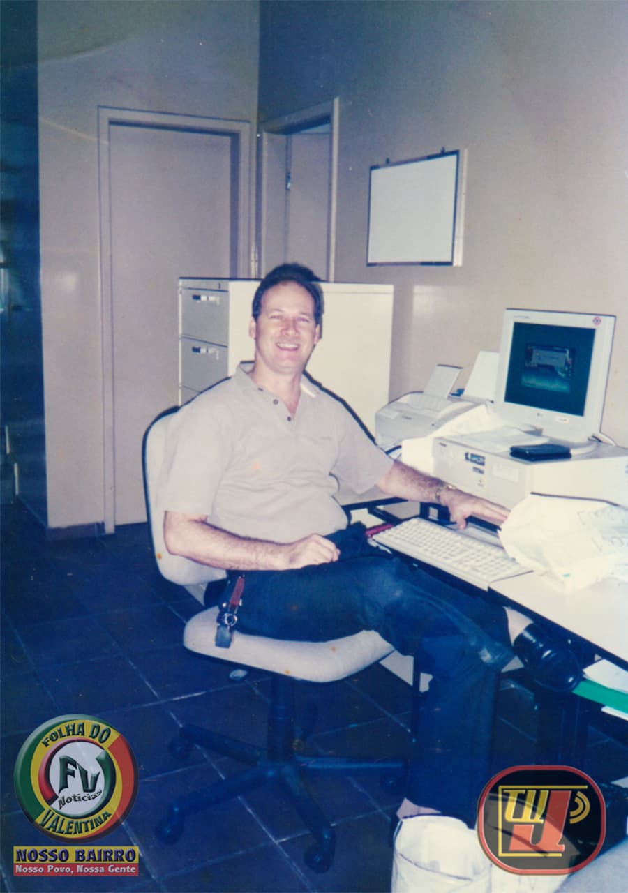 Fotografia colorida de época registrada em ambiente interno de escritório por volta do ano 2000 ou 2001, mostrando Petrúcio Prado em sua mesa de trabalho no Banco Real. Ele está sentado em uma cadeira giratória cinza, sorrindo para a câmera, vestindo uma camisa polo bege clara e calça escura. Sua mão esquerda repousa sobre um teclado de computador de modelo antigo (CRT de tubo) posicionado em uma mesa de escritório branca ao lado de uma impressora e papéis. Ao fundo, vê-se um arquivo de aço bege com gavetas, portas de escritórios e um pequeno quadro branco fixado na parede. No canto inferior esquerdo está o selo circular "Folha do Valentina" e, no canto inferior direito, o logotipo da TV Jampa.