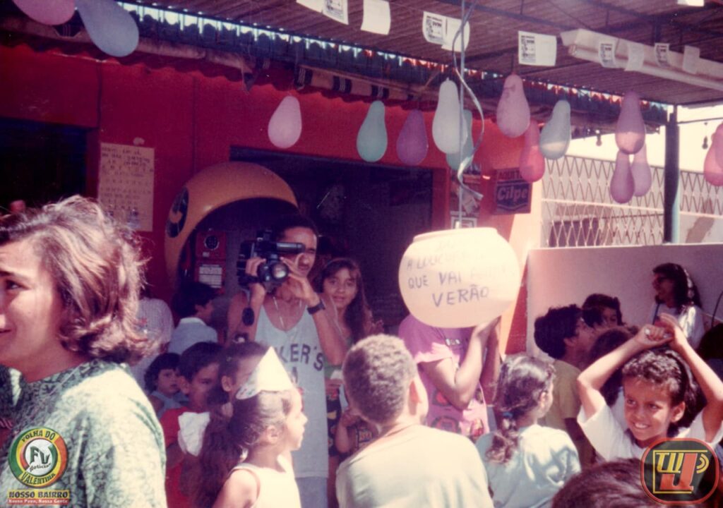 Fotografia colorida de época, tirada ao ar livre nos anos 90 sob a sombra de uma árvore, registrando uma entrega de prêmios durante o evento beneficente "Prado Picolé e Dudu" no Natal de 1994. No centro da imagem, Petrúcio Prado jovem, sorridente, veste uma camiseta regata cinza com detalhes vermelhos e estende as mãos entregando um brinde. À sua esquerda, em primeiro plano e de perfil, aparece uma pessoa fantasiada de Papai Noel com gorro e barba branca. Ao fundo e à direita, várias crianças e adultos observam a cena com alegria. No canto inferior esquerdo está o selo "Folha do Valentina" e, no canto inferior direito, o logotipo clássico da TV Jampa.