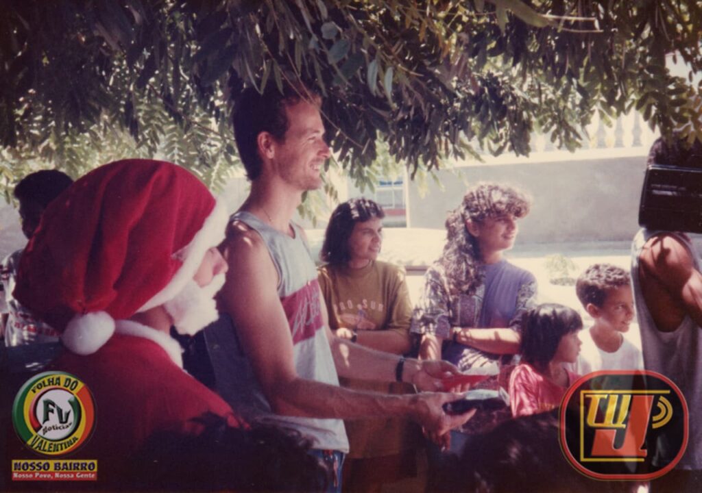 Fotografia colorida de época, tirada ao ar livre nos anos 90 sob a sombra de uma árvore, registrando uma entrega de prêmios durante o evento beneficente "Prado Picolé e Dudu" na época de natal em 1994. No centro da imagem, Petrúcio Prado jovem, sorridente, veste uma camiseta regata cinza com detalhes vermelhos e estende as mãos entregando um brinde. À sua esquerda, em primeiro plano e de perfil, aparece uma pessoa fantasiada de Papai Noel com gorro e barba branca. Ao fundo e à direita, várias crianças e adultos observam a cena com alegria. No canto inferior esquerdo está o selo "Folha do Valentina" e, no canto inferior direito, o logotipo clássico da TV Jampa.
