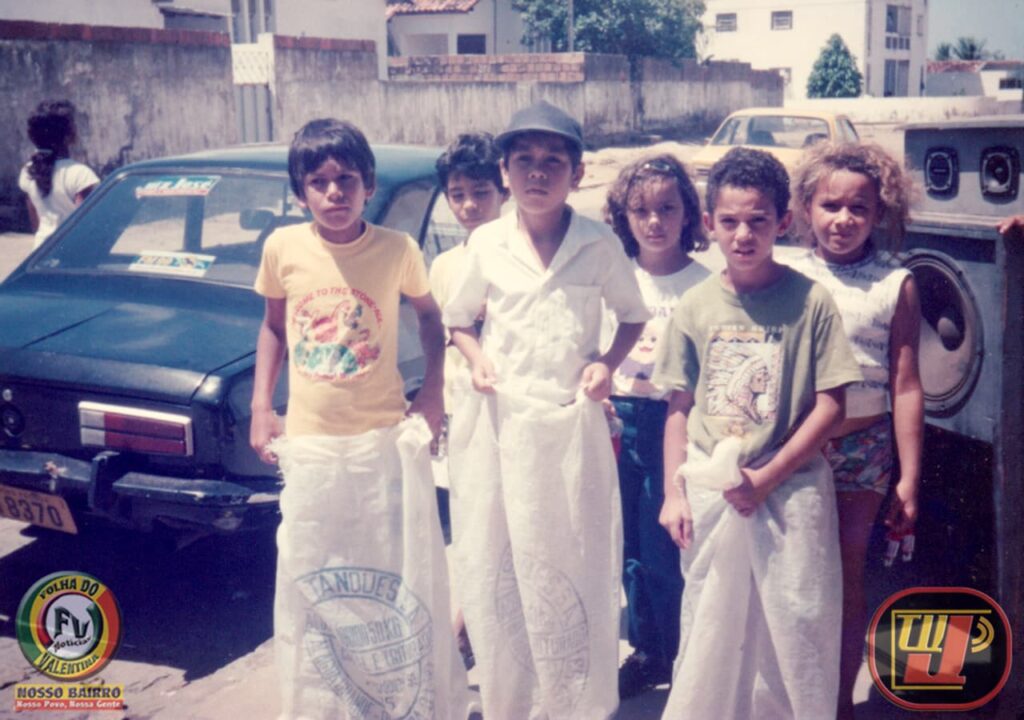 Fotografia colorida de época registrada ao ar livre em uma rua residencial no Natal de 1994, mostrando crianças preparadas para uma corrida de sacos na festividade "Prado Picolé Dudu". Em primeiro plano, três meninos estão posicionados lado a lado com as pernas dentro de grandes sacos de ráfia branca (sacos de linhagem usados para transporte de mercadorias), segurando as bordas com as mãos. Atrás deles, outras três crianças observam e aguardam o início da brincadeira. Ao fundo, à esquerda, há um carro antigo de cor escura estacionado e, à direita, uma grande caixa de som antiga com alto-falantes aparentes na calçada. No canto inferior esquerdo está o selo circular "Folha do Valentina" e, no canto inferior direito, o logotipo da TV Jampa.