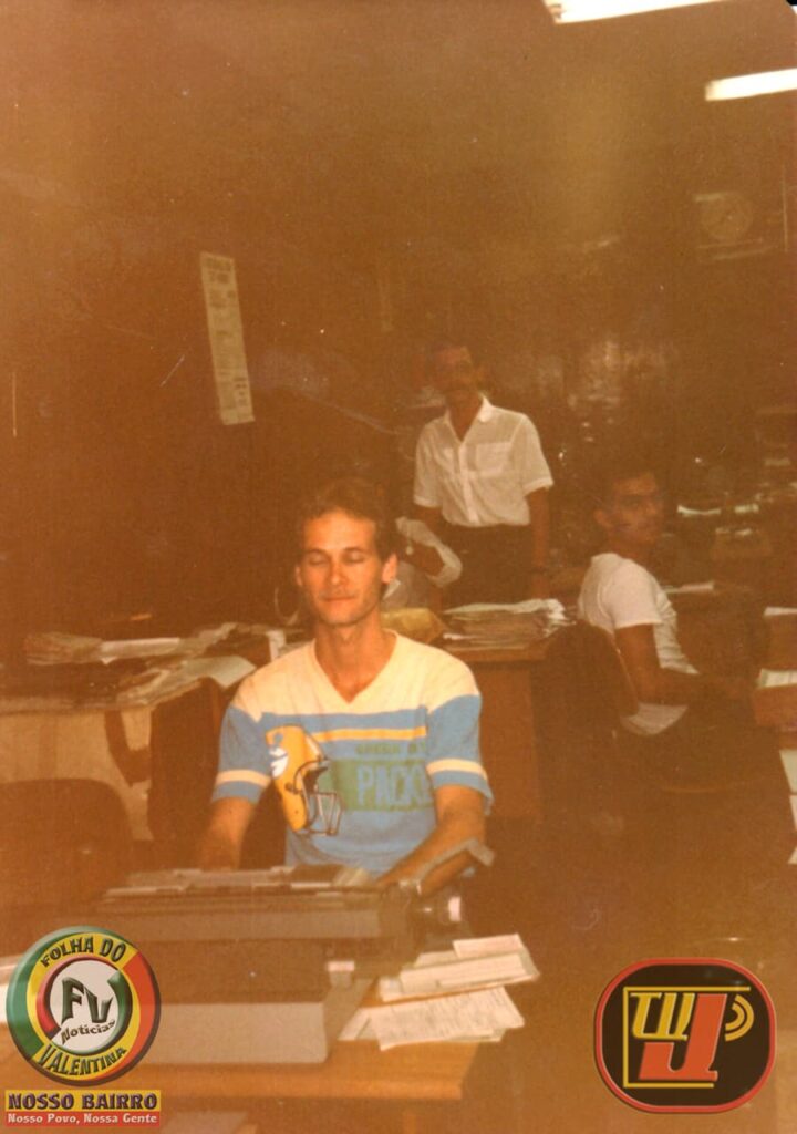 Uma foto vintage colorida, com um tom sépia característico dos anos 80, mostrando Petrucio Prado em primeiro plano, sentado e operando uma máquina de datilografia branca e cinza. Ele veste uma camiseta branca com faixas azul-claras e amarelas e uma estampa de desenho animado. Seus olhos estão fechados e ele parece focado no trabalho. Ao fundo, em um ambiente de escritório em banco, dois outros homens são visíveis: um em pé, sorrindo e olhando para a câmera, e outro sentado em uma mesa adjacente, também de frente para a câmera. Pilhas de documentos e arquivos estão espalhadas pelas mesas e prateleiras ao fundo. No canto inferior esquerdo, há um logotipo circular com um 'F' e um 'V' entrelaçados e a inscrição 'FOLHA DO VALENTINA' e 'NOSSO BAIRRO', enquanto o canto inferior direito apresenta um logotipo circular com as letras 'T', 'W', 'P' e 'C' estilizadas.