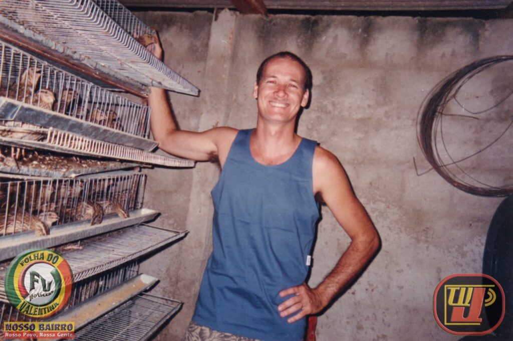 Foto colorida de um homem sorridente de regata azul clara, em pé em um criatório. Ele está posicionado no centro, com a mão esquerda na cintura e o braço direito erguido, tocando suavemente a lateral de uma grande estrutura de gaiolas aramadas à esquerda. As gaiolas estão organizadas em andares e abrigam dezenas de codornas. O fundo é uma parede de concreto simples e há um rolo de arame pendurado à direita. No canto inferior esquerdo, estão os logotipos "Folha do Bairro Notícias - Valentina" e "NOSSO BAIRRO". No canto inferior direito, o logotipo da "TV Jampa".