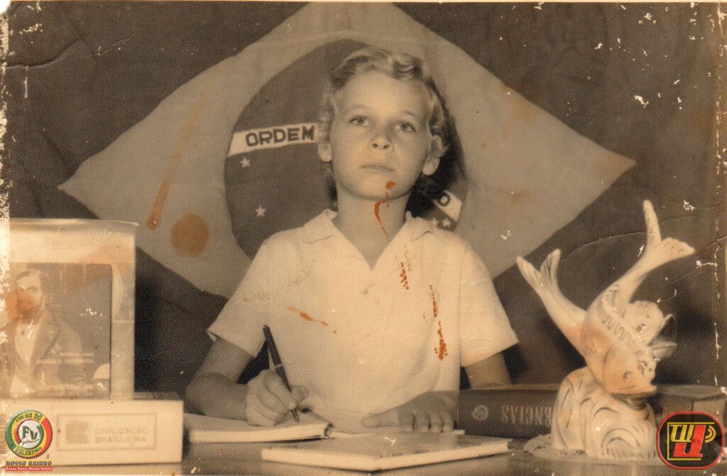 Fotografia em tom sépia de um menino loiro de 6 anos sentado em uma mesa escolar em 1966. Ao fundo, está a bandeira do Brasil. O menino veste camisa polo clara e segura uma caneta sobre um caderno aberto. Sobre a mesa, há livros didáticos (um deles com o título "Civilização Brasileira" e outro escrito "Ciências" de cabeça para baixo) e uma escultura decorativa de peixes saltando. A imagem possui marcas do tempo, manchas alaranjadas e logotipos modernos nos cantos inferiores ("Folha do Valentina" à esquerda e "TV Jampa" à direita).