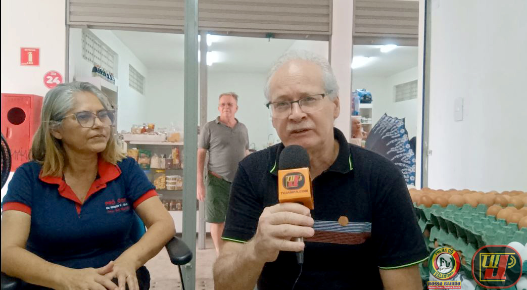 Foto colorida de formato horizontal, em plano médio, tirada durante uma entrevista no dia 16 de julho na Pró Ovos. No centro, o jornalista Petrúcio Prado, de óculos e camisa polo preta com listras brancas e vermelhas, fala ao microfone da TV Jampa. À esquerda, Maria Verônica, também de óculos e com o uniforme da Pró Ovos, uma camisa polo azul com gola vermelha, escuta atentamente. Ao fundo, vê-se a loja da Pró Ovos, com prateleiras de mantimentos e um homem de camisa cinza em pé. No canto inferior direito, estão os logotipos da TV Jampa e do Portal Folha do Valentina. A imagem tem a referência verbatim "Pró Ovos - Marie Verônica - tvjampa petrucio prado portal folha do valentina 8-.jpg".