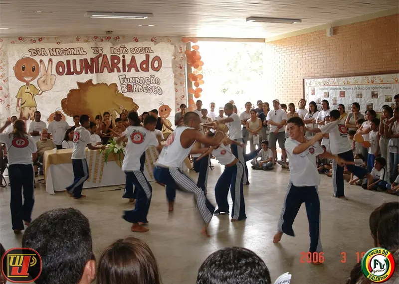 Grupo de jovens realiza apresentação cultural de capoeira e maculelê em um grande salão durante o IV Dia Nacional de Ação Voluntária Fundação Bradesco, cercados por uma plateia de espectadores.
