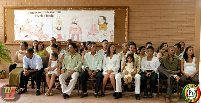 Diversos casais de noivos e suas famílias sentados em fileiras de cadeiras em um auditório com parede de tijolos à vista, aguardando o início da cerimônia do casamento coletivo na Fundação Bradesco.