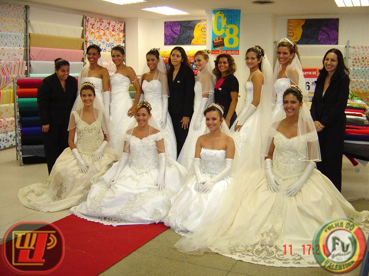Fotografia colorida em plano geral reunindo modelos e organizadoras no setor de tecidos de uma loja durante a 3ª Expo Noivas Esplanada. As modelos vestem luxuosos vestidos de noiva brancos com tiaras, véus e luvas longas; quatro estão sentadas na passarela vermelha e as demais posam em pé ao fundo junto à estilista Cristiane Lima (Mylena Noivas) e equipe. No canto inferior esquerdo está a marca da TV Jampa e no canto inferior direito a marca da Folha do Valentina com a data impressa em vermelho "11 17:21".