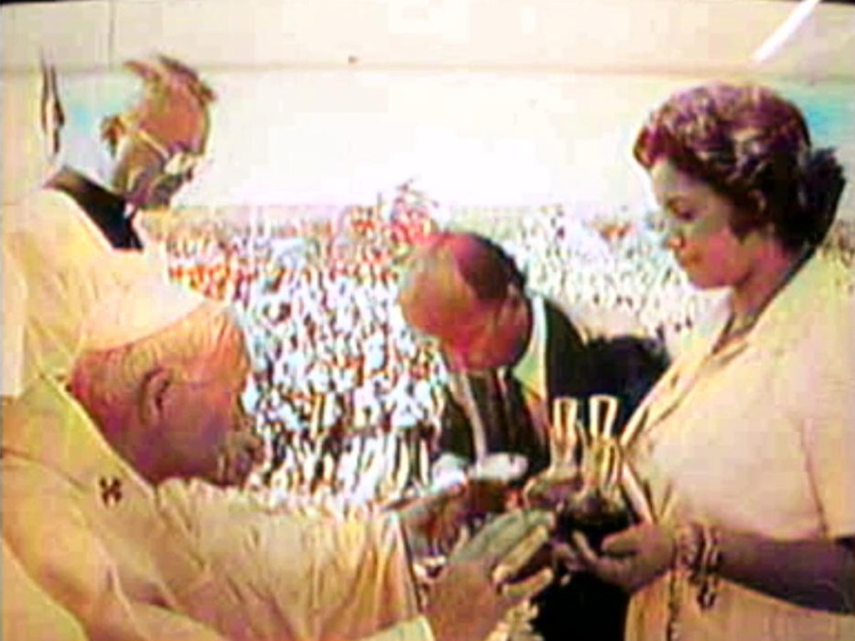 Fotografia colorida antiga mostrando o Papa João Paulo II, visto de perfil à esquerda com túnica branca, recebendo a apresentação das galhetas com água e vinho de uma mulher e um homem durante uma celebração religiosa campal, com uma grande multidão ao fundo.