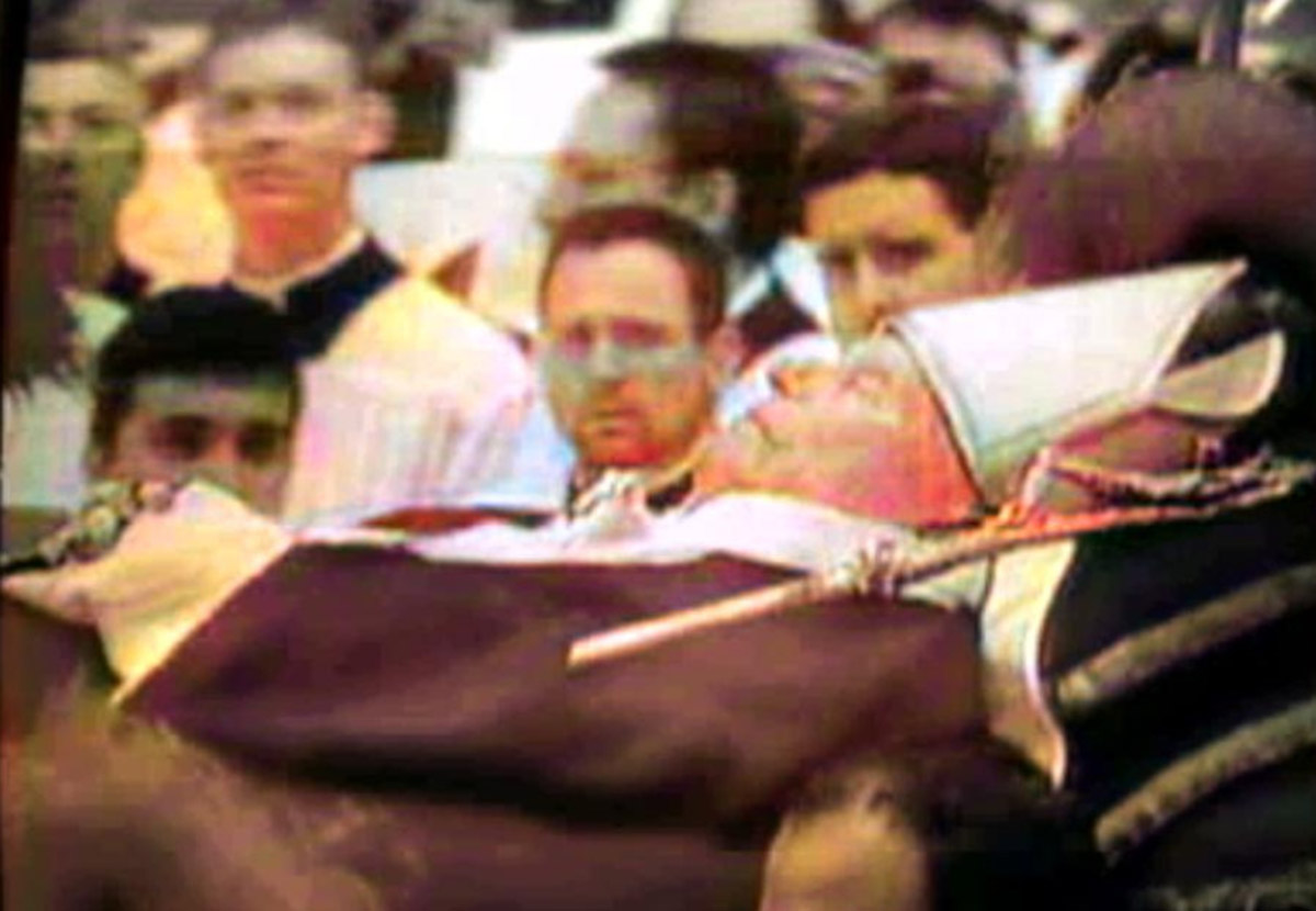 Fotografia colorida antiga, com baixa resolução e granulada, capturada de uma tela de vídeo. A imagem mostra o Papa João Paulo II em repouso após sua morte, deitado sobre um esquife. Ele está vestido com vestes papais brancas e vermelhas e usa uma mitra branca na cabeça. Seu rosto está visível no centro-direito, com olhos fechados. Ao fundo, uma multidão desfocada de pessoas de luto e observadores, incluindo um homem de óculos no centro e outras figuras religiosas em vestes escuras. O ambiente parece ser um grande espaço interno. A qualidade da imagem sugere uma gravação de arquivo analógico da época.