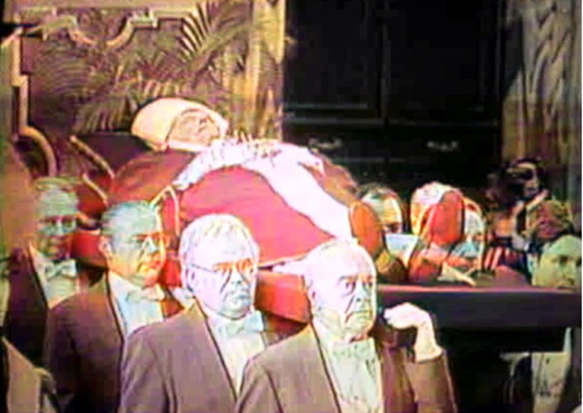 Uma captura de tela granulada, colorida e de baixa resolução, obtida de um vídeo analógico antigo, mostra um cortejo fúnebre para o Papa João Paulo II. O corpo do falecido Papa está em repouso sobre uma plataforma, vestido com paramentos papais vermelhos e brancos completos, incluindo a mitra e casula vermelha. Suas mãos estão cruzadas sobre o peito. No primeiro plano, vários homens mais velhos em ternos pretos formais e gravatas borboleta pretas, com expressões sombrias, carregam a plataforma sobre os ombros. Dois homens brancos de óculos estão mais próximos da câmera à esquerda, enquanto outros estão à direita. O fundo é escuro e indefinido, com alguns detalhes arquitetônicos e folhagens que sugerem o interior da Basílica de São Pedro ou um espaço semelhante durante o funeral. A imagem tem a qualidade visual de uma gravação de vídeo VHS antiga, com distorção de cores e ruído.