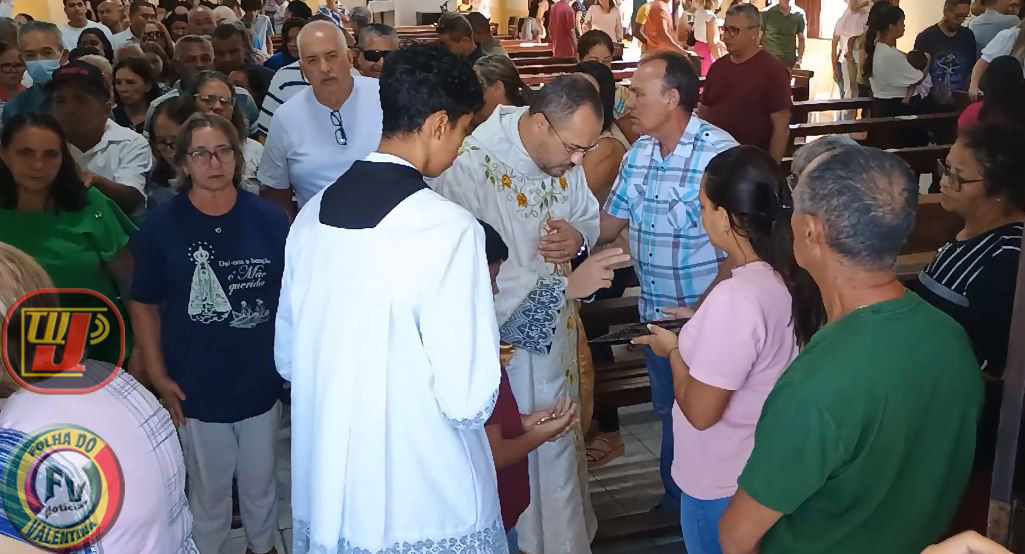 Celebrando a Assunção da Virgem Maria, o Padre Luiz Carlos abençoa fiéis no corredor central da Paróquia Nossa Senhora Aparecida rodeado por coroinhas e comunidade. Imagem com marcas d'água da TV JAMPA e Folha do Valentina Notícias.