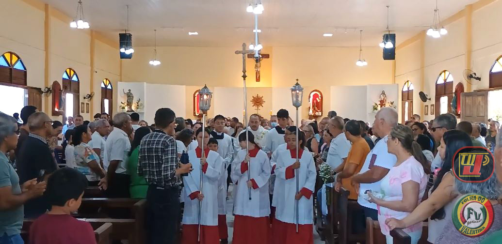 Visão do corredor central da Paróquia Nossa Senhora Aparecida lotada de fiéis durante a procissão de entrada, com coroinhas vestindo túnicas brancas e vermelhas segurando a cruz procissional e lanternas, seguidos pelo Padre Luiz Carlos ao fundo. A imagem possui as marcas d'água da TV JAMPA e Folha do Valentina Notícias.