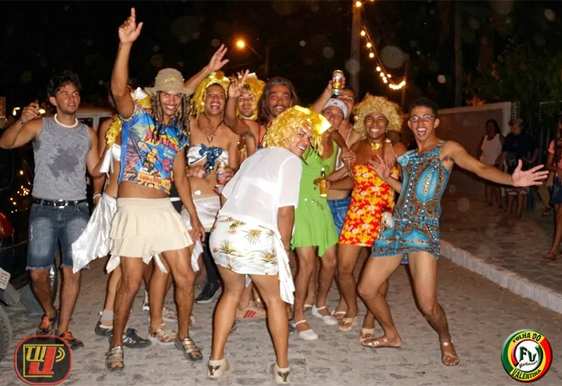Grupo de homens fantasiados de mulher sorriem e fazem brincadeiras no bloco Peruas do Valentina. Eles vestem saias, vestidos coloridos e perucas loiras, celebrando em uma rua de paralelepípedos durante a noite. Ao fundo, há cordões de luzes decorativas e outras pessoas assistindo à festa.