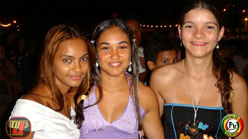 Três jovens foliãs sorriem lado a lado no bloco Peruas do Valentina. A jovem à esquerda tem cabelos claros e blusa branca ciganinha; a do centro usa blusa roxa com detalhes brilhantes; e a da direita veste um top preto com estampa de borboletas. Ao fundo, vê-se o público da festa noturna e dois logotipos circulares nos cantos inferiore