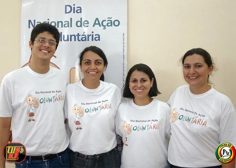 Quatro professores e funcionários sorriem abraçados vestindo a camiseta oficial do evento em frente a um banner informativo durante o IV Dia Nacional de Ação Voluntária Fundação Bradesco.